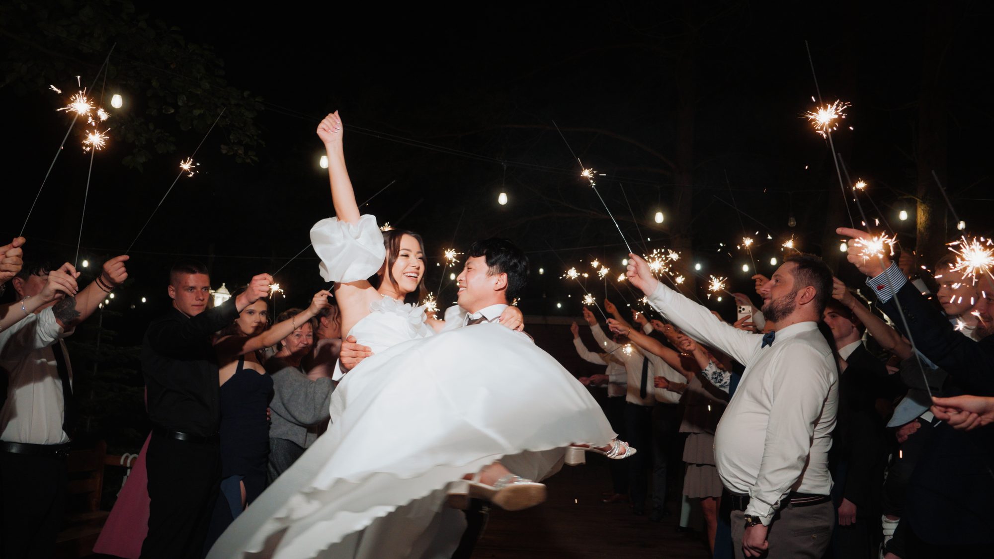 Pan młody unosi pannę młodą w szpalerze gości z zimnymi ogniami, wesele w plenerze w Woodland Wedding w Starych Żukowicach